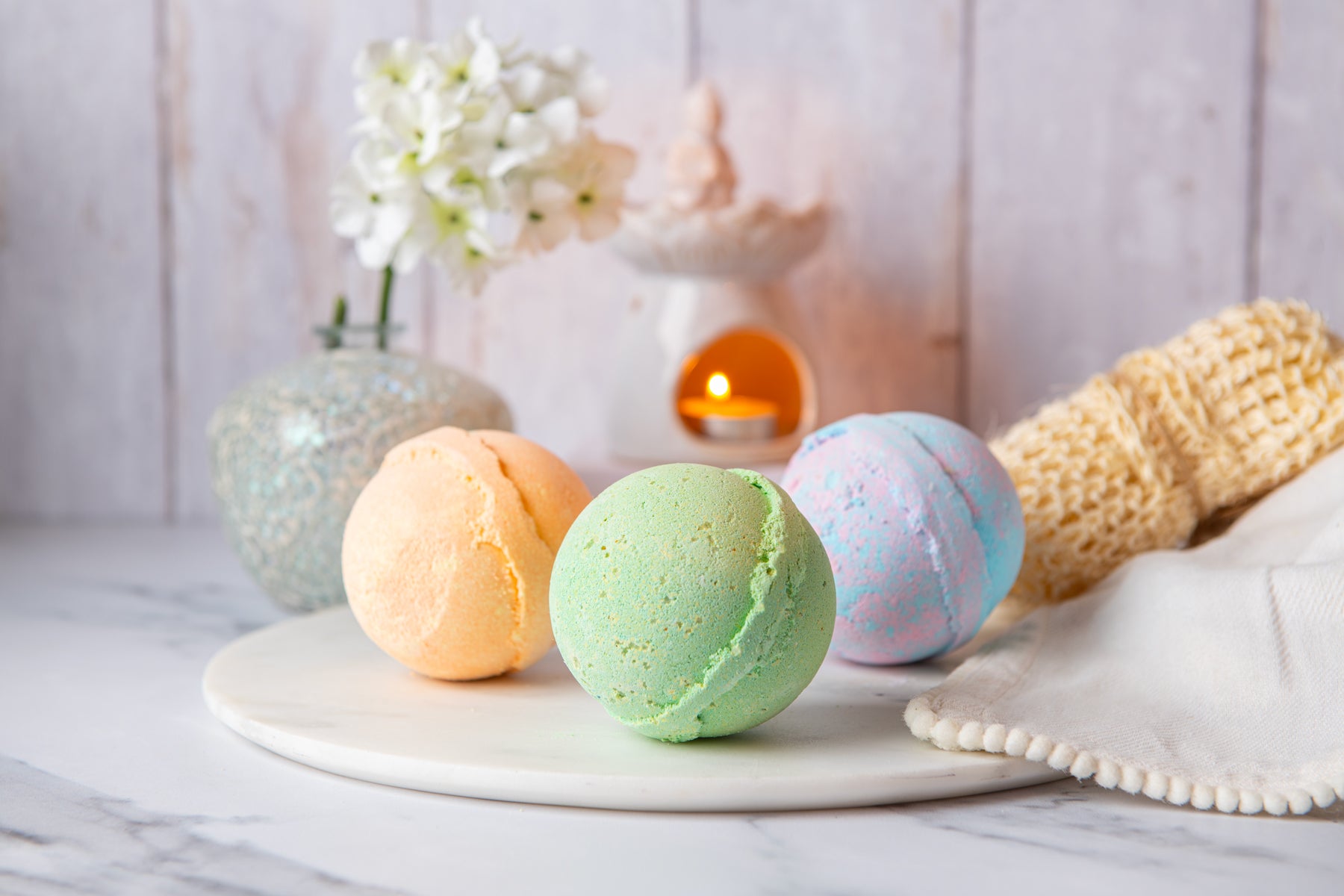 Three colorful bath bombs on a marble surface with a candle and flowers in the background.