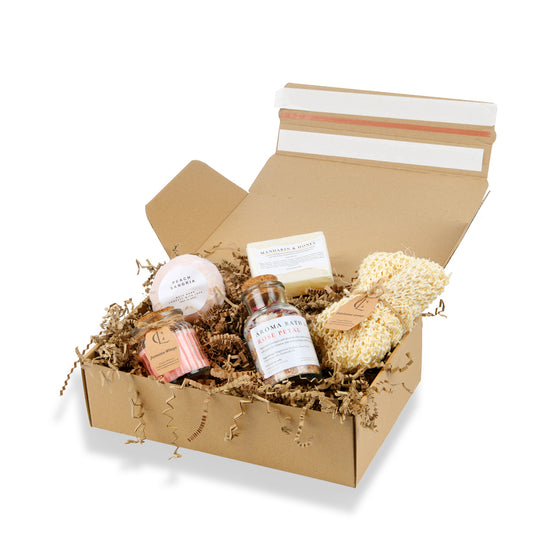 Open cardboard box with bath products including a loofah, soap, and bath bombs on a white background