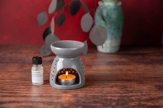 Gray ceramic oil burner with a lit candle on a wooden surface, next to a small bottle of essential oil.