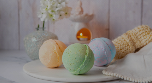 Three colorful bath bombs on a white surface with a blurred background featuring a candle and flowers.
